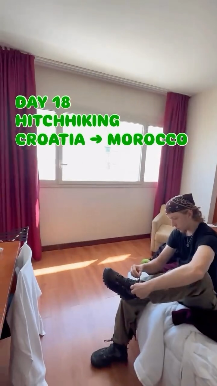 Lacing a boot on the edge of a bed in a bare hotel room, bandana on, red curtains pulled back from a blown-out window.