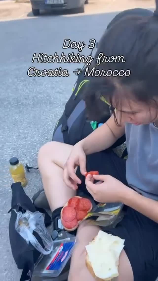 Sitting on the kerb beside a loaded pack, building a sandwich: tomato sliced onto bread, a bottle of juice and a hunk of cheese on the ground.